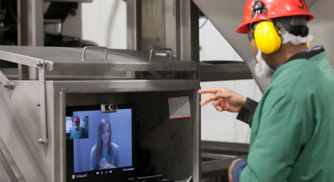 A hard hat worker stands within a factory, video calling with another individual, signaling something.