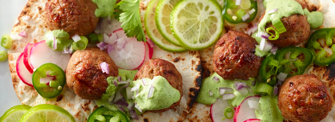 A collection of meatballs, green sauce, sliced limes, radishes, onions, and peppers lay atop toasted flat bread.