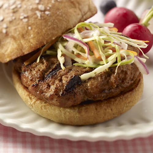 A delicious turkey burger, topped with homemade coleslaw, served with a side of vegetables on a white plate, with a pink tablecloth.