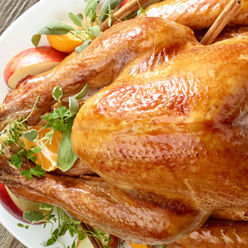 An apple brined whole turkey served with herb sprigs, cinnamon sticks, and fruit, served on a white plate on a wooden table.