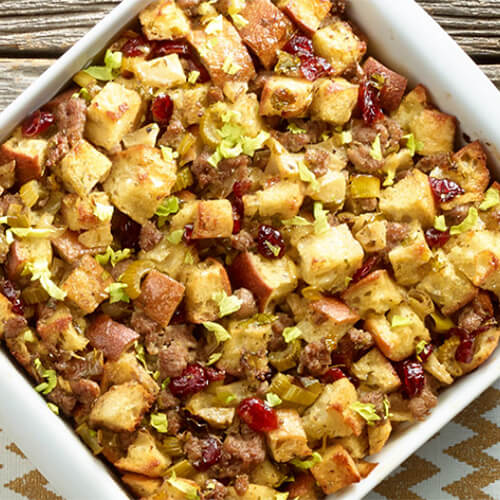 Stuffing filled with apple, turkey sausage, and dried cranberries, served in a white dish on a golden tablecloth and a wooden table.