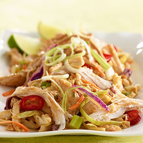 Vegetables, coleslaw mix, and turkey combine with crispy noodles to create an Asian-inspired salad, served ona white plate garnished with lime wedges, atop a green cloth.