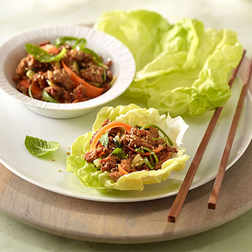 Crisp lettuce wraps on a white plate filled with cucumber, ground turkey, and spicy pepper sauces, served in a white bowl, on a wooden plate.