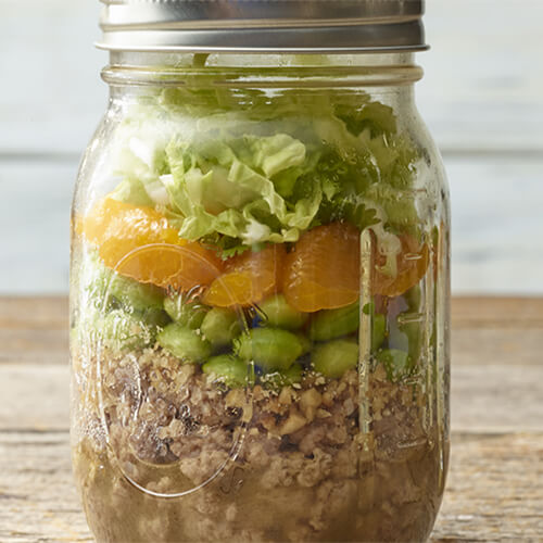 A ginger sesame dressing, turkey, cashews, edamame, mandarin oranges, cabbage and cilantro all served in a mason jar on a wooden table.