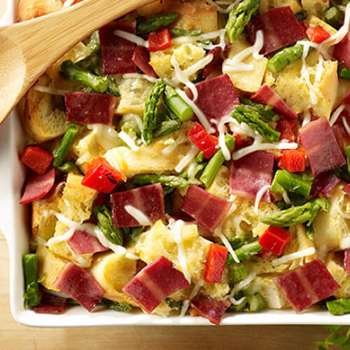 A hearty strata made from egg, turkey, bacon, and asparagus within a casserole tray, on a wooden plate.