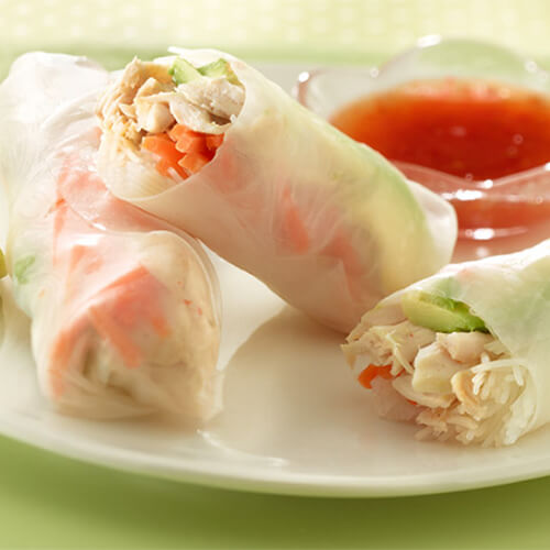 Shredded turkey, avocado and noodles rolled in rice paper and served with a sweet chili sauce, on a white plate garnished with a lime wedge, on a green cloth.