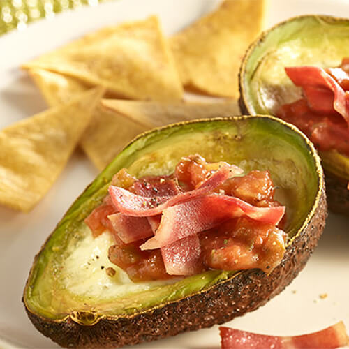 Avacado halves filled with crisp turkey bacon and baked eggs, served on a white plate with tortilla chips.
