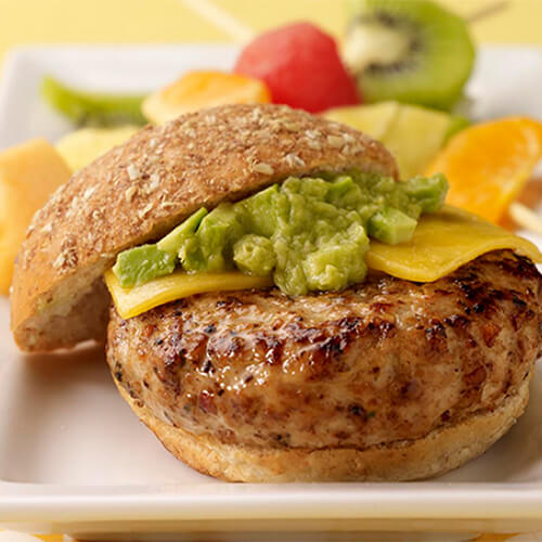 A burger with a patty made from a mix of applesauce and turkey, topped with avocado and cheese, served with a side of fruits kabobbed, on a white tray, on a yellow floral table.