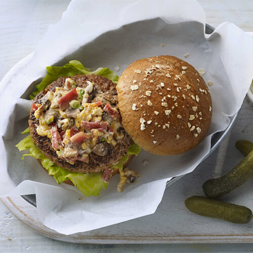 Homemade burgers made from ground turkey and spices, topped with lettuce, cheese, bacon, mushrooms, and mayonaise, served on a brioche bun, with a side of pickles and mayonaise, served on a wooden table.