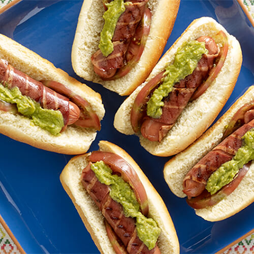 Bacon wrapped turkey franks served with guacamole and tomatoes, on a blue plate atop a painted tablecloth.