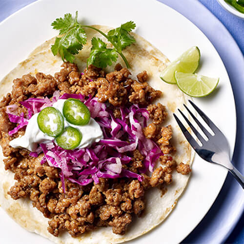 A hearty portion of lean turkey in a taco, topped with a jicama and cayenne pepper-infused slaw, served on a white plate on top of a blue plate atop a blue napkin.