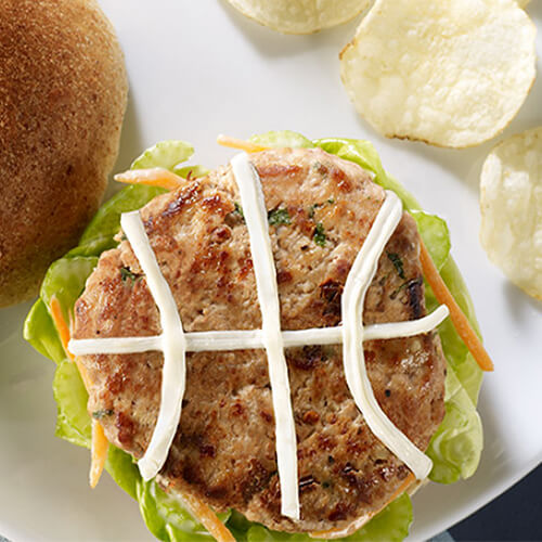 Hot turkey patties decorated with cheese strings, and topped with a blue cheese dressing and vegetables, served on a whole grain bun with a white plate, with a side of potato chips.