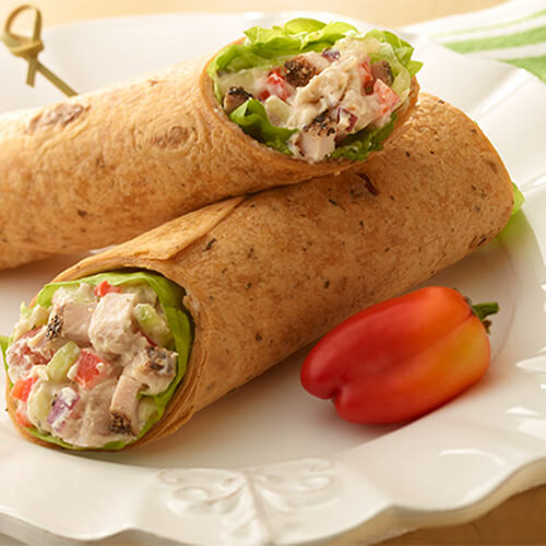 A wrap filled crunchy veggies, a creamy ranch spread and a spicy kick from diced cracked pepper turkey, served with a red bell pepper on a white plate.