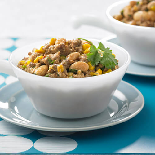 Meaty, smoky, chili filled with ground turkey, corn, beans, broth, chipotle peppers and adobo sauce served in a white small soup bowl on a blue tablecloth.