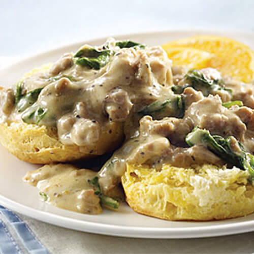 Freshly baked biscuits smothered in a savory spinach and turkey sausage gravy, served on a white plate garnished with an orange slice.