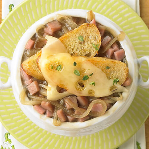 A hearty portion of turkey ham, beef broth, and golden brown onions, on a white bowl, topped with cheesy French bread, on a green plate.