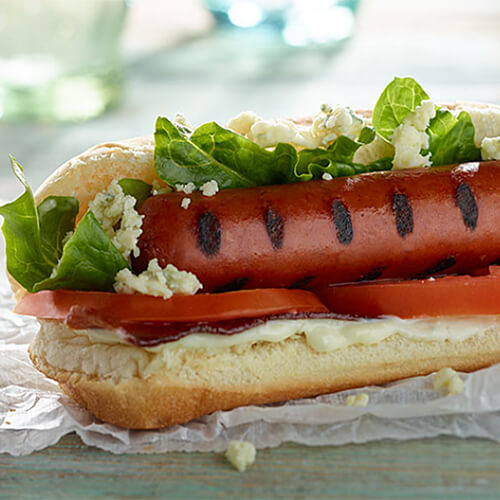 A frank filled with blue cheese dressing, bacon, tomatoes, and lettuce, served in a hotdog bun on parchment paper.