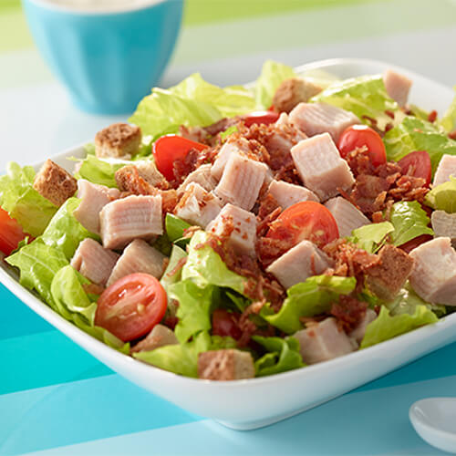 A salad filled with crispy turkey bacon, cubed roast turkey breast and fresh cherry tomatoes on a bed of romaine lettuce, served in a white bowl atop a blue colorful table mat.