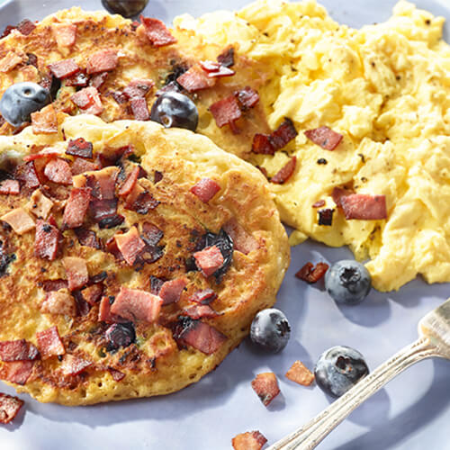 Two pancakes topped with blueberries and bacon, next to a side of eggs on a blue plate.