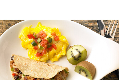 A great portion of eggs topped with peppers, served with a hearty portion of peppers, beans, and ground turkey in a tortilla, with a side of kiwi wedges, on a white plate atop a wooden table.