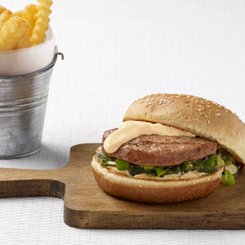 A grilled turkey burger topped with grilled onions and peppers, a spicy aioli and a melted cheese sauce, served on a wooden board and a small metal bucket filled with crinkle cut french fries.