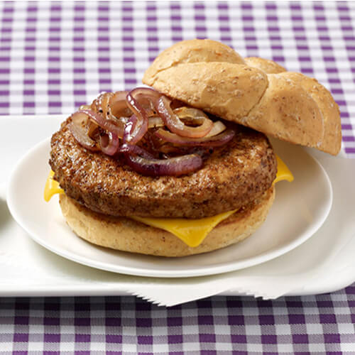 A seasoned and grilled turkey burger topped with sauteed onions, and melted cheese, served on white plate garnished with pickles with a purple checkered table cloth.