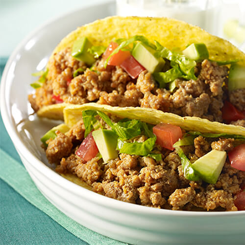 Flavorful tacos made with buffalo-style turkey, fresh vegetables and blue cheese dressing, served with a side of milk and celery on a white plate atop a colorful blue tablecloth.