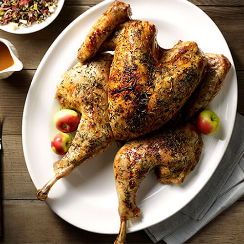 A gorgeous herbed turkey served with miniature apples, served with homemade apple sherry gravy and wild rice stuffing, served on a white plate atop a wooden table.