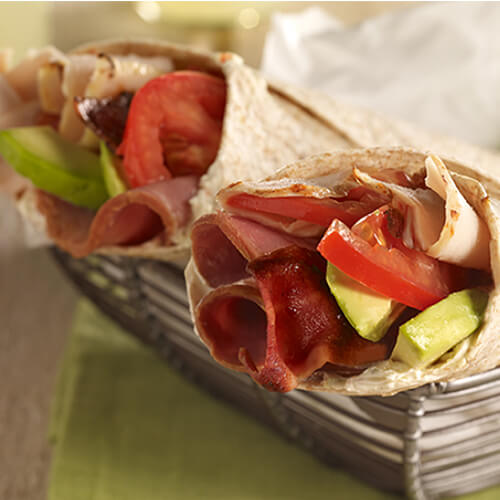 2 crispy flour wraps filled with turkey breast, turkey ham, creamy avocado, and fresh tomatoes in a woven bowl on a wooden table.