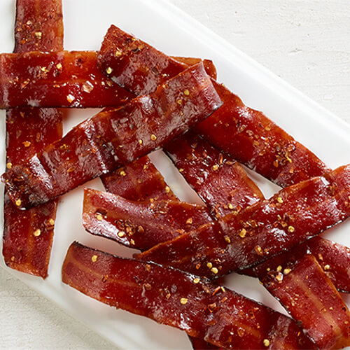 Bacon slices baked and dipped in maple syrup, sprinkled with brown sugar and crushed red pepper flakes, served on a white plate atop a wooden table.