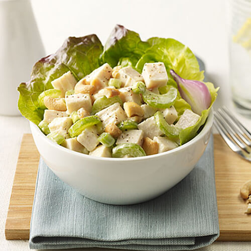 A hearty portion of turkey roast, celery, onions, and cashew pieces served on a white bowl with a curry mayo atop a blue cloth and wooden table.