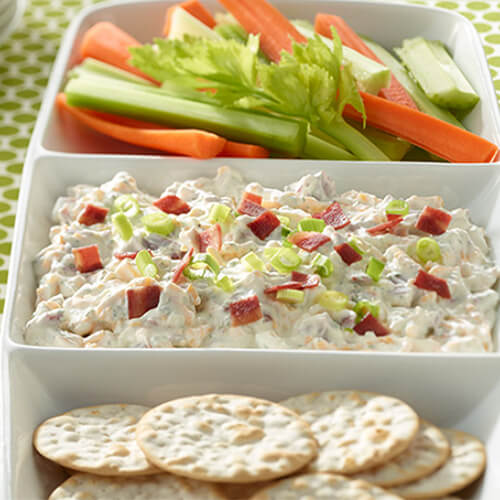 A combination of sour cream, ranch, cheddar, and crispy turkey bacon, served with sides of vegetables and crackers, served on a white platter on a green tablecloth.