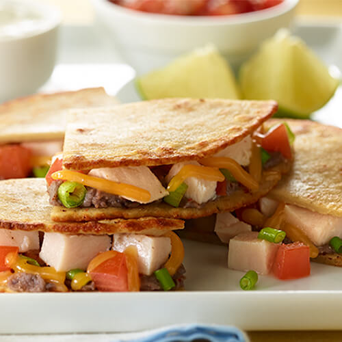 A lovely quesadilla filled with Melted cheese, deli turkey, refried beans, fresh tomatoes and green onions, served on a what plat with a side of salsa, sour cream, and lime wedges.