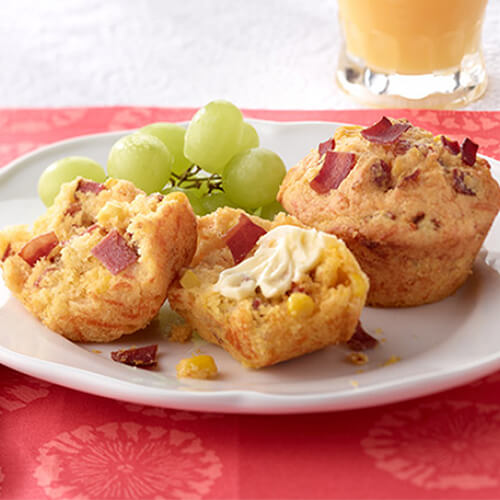 Savory corn muffins filled with crisp turkey bacon, melted Mexican cheese and delectable honey butter, served with grapes on a white plate, atop a red tablecloth.