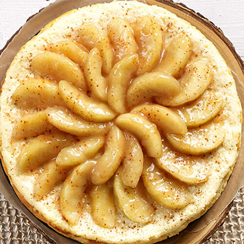 A delectable cheesecake topped with melted white chocolate and cinnamon apples, served in a pie tray with parchment paper, served in a wired table mat.