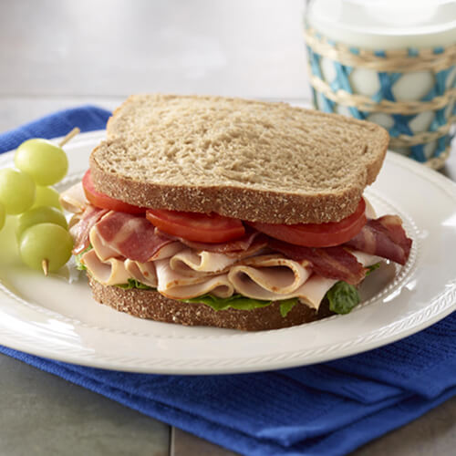 A classic sandwich piled with lettuce, tomato, lean turkey bacon, and herb roasted turkey breast, served with skewered grapes on a white plate with a glass of milk on a blue cloth.