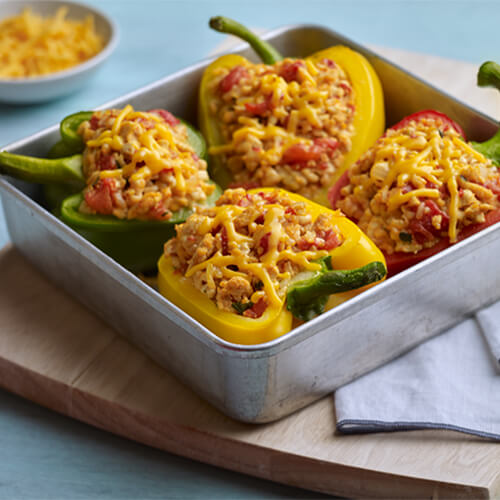 Colorful bell peppers stuffed with lean ground turkey, rice, tomatoes, cheese, and served in a metal baking dish atop a wooden board.