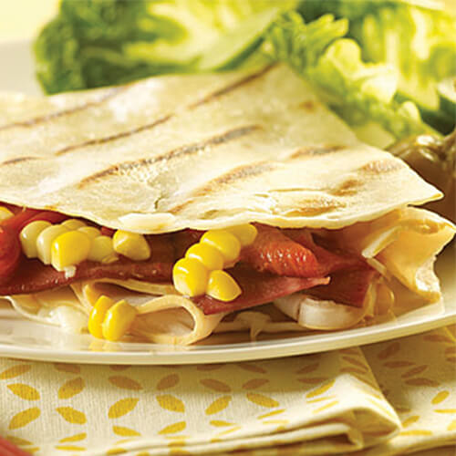 Hearty quesadillas loaded with freshly grilled corn, roasted red pepper, flavorful turkey and turkey bacon, served on a white plate atop a yellow floral cloth.