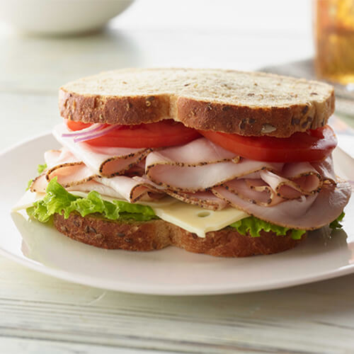 A sandwich topped with cracked pepper turkey, swiss cheese, lettuce, tomato, and onions, on a white plate atop a white table.