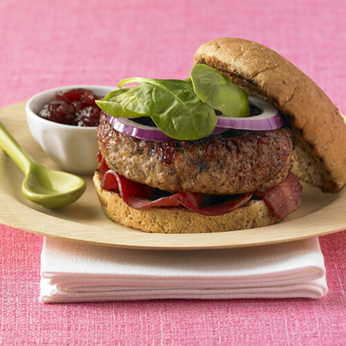 A grill-fired turkey burger topped with lean turkey bacon and spinach served with a flavorful cranberry sauce on a wooden plate with a pink tablecloth.