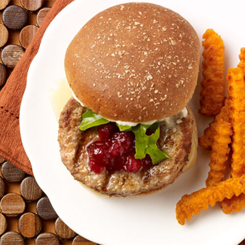 A savory turkey burger topped with horseradish, Swiss cheese, sweet cranberry sauce, served on a lean hamburger bun, served with a side of sweet potato fries, on a white plate, atop a wooden buttoned table mat.