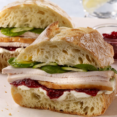 A cibatta sandwich filled with roast turkey and cranberry sauce, fresh spinach, and crunchy cucumbers, served on parchment paper.