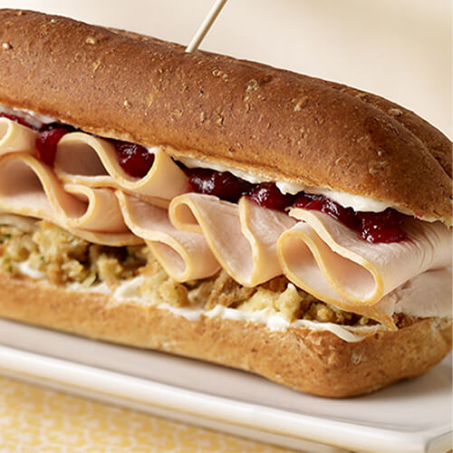 Juicy turkey, stuffing, and cranberry relish layered on a hoagie roll, on a white plate, on a yellow patterned cloth.