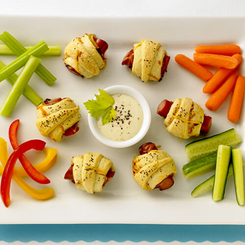 Crescent rolls wrapped around JENNIE-O® turkey franks served with vegetable with a honey mustard sauce, served on a white tray on a blue place mat with a white background.