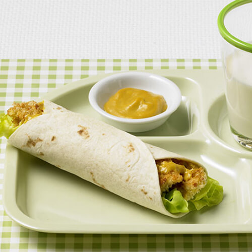 A warm tortilla filled honey mustard, lettuce and crunchy baked turkey, served with sides of grapes, honey mustard, and a glass half-full of milk in a green tray with dividers, atop a green checkered tablecloth.