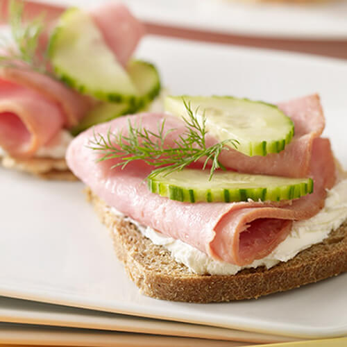 Slices of rye bread topped with cream cheese, turkey ham, cucumber, and dill atop white plates, with a pink and yellow background.