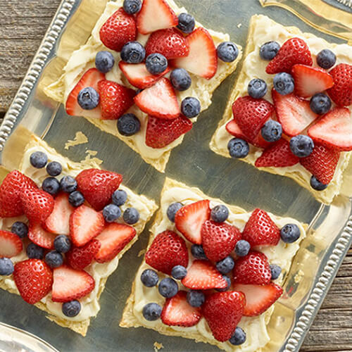 A dessert made from puff pastry dough, marscapone cheese and topped with a healthy portion of berries served on a metal tray atop a wooden table.