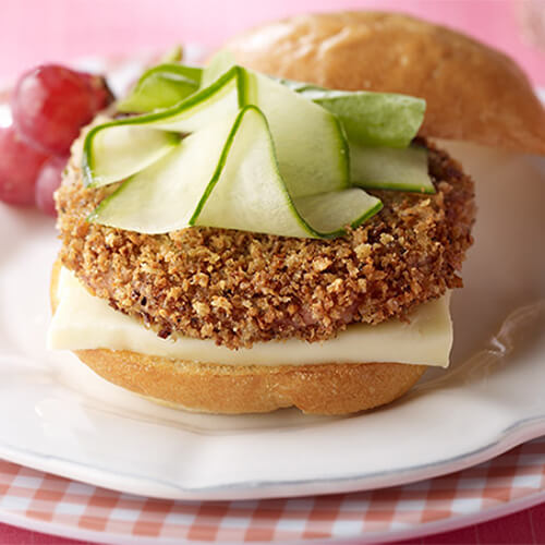 A crispy breaded turkey burger topped with soft brie cheese and crunchy cucumber garnished with tomatoes, served on white and checkered plates, and on a pink tablecloth.