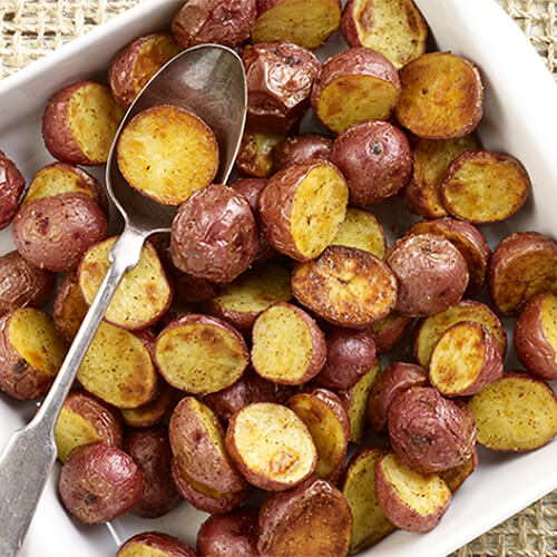 Delectable roasted potatoes tossed with with olive oil, Creole seasoning and salt, served in a white bowl on a wired tablecloth.