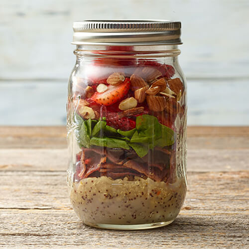 A lemon-poppyseed dressing, quinoa, turkey bacon, spinach, strawberries and almonds all served in a mason jar on a wooden table.